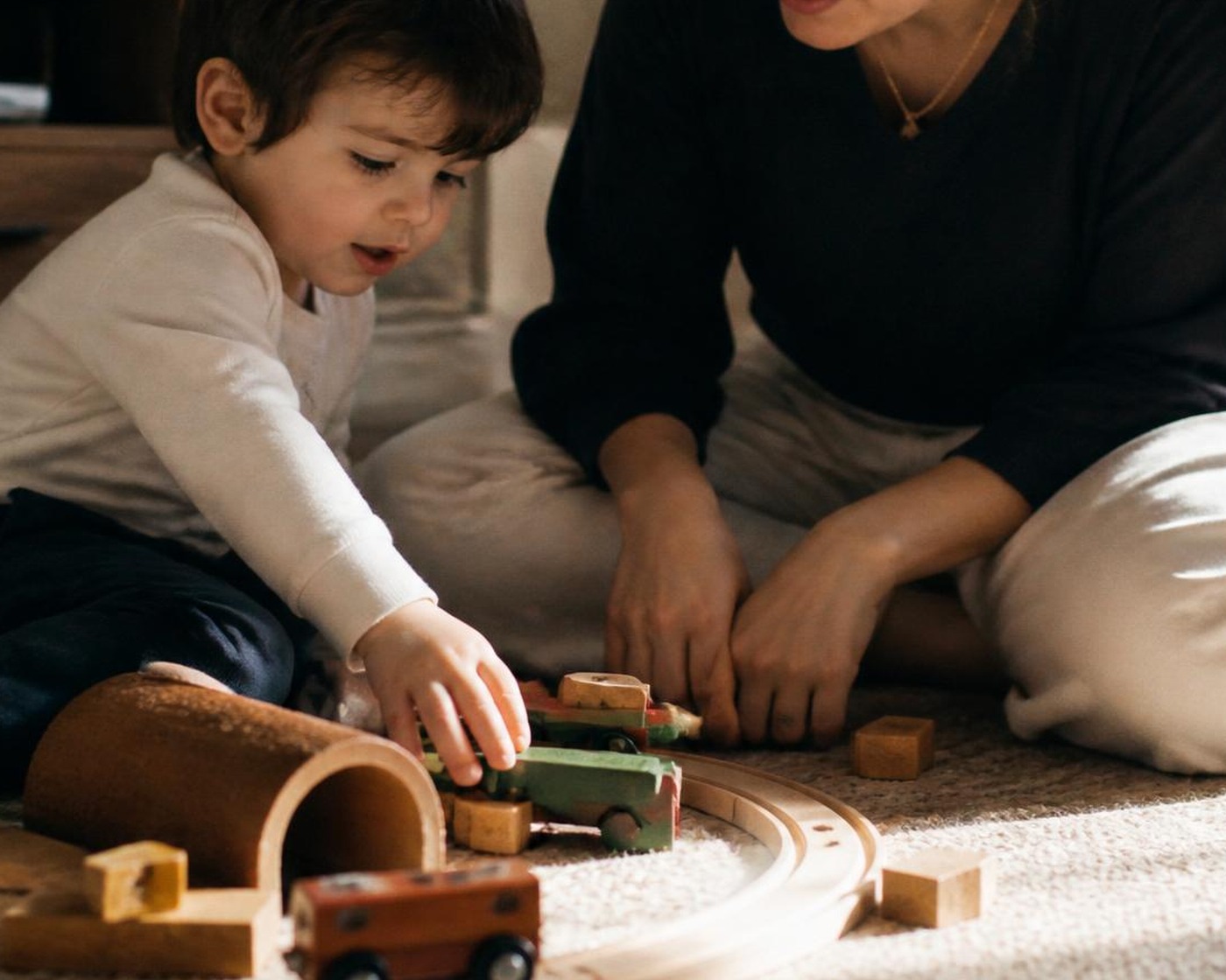 Toddler and caregiver kneeling on the rug together, guiding a wooden train around its track